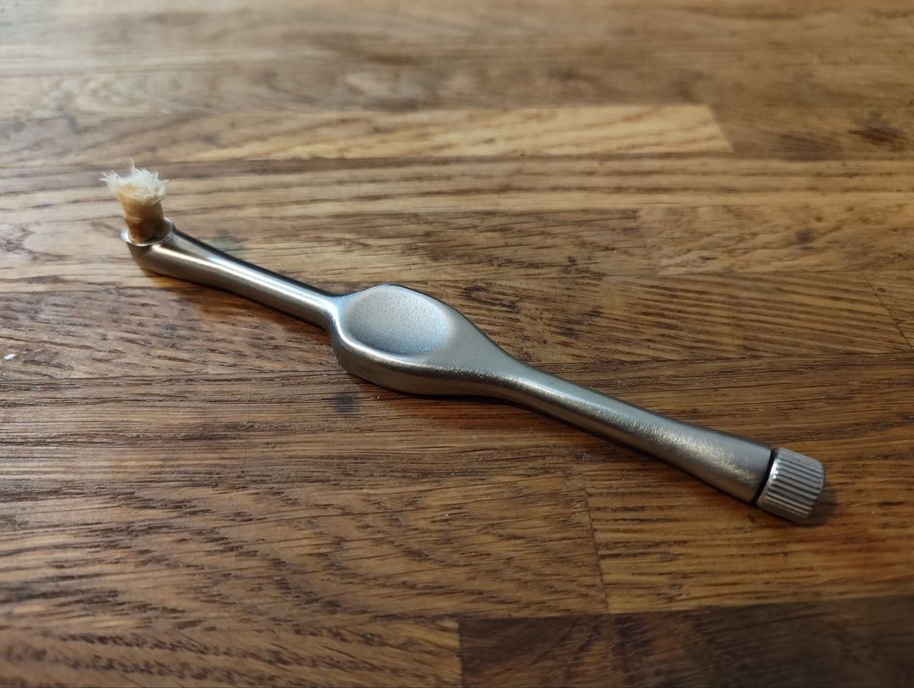 Der Zahnputzhobel-Prototyp: ein gebürsteter Edelstahlgriff mit einer eingeklemmten Miswak-Spitze an einem Ende und einer geriffelten Spannmutter am anderen, auf einem Eichentisch liegend.